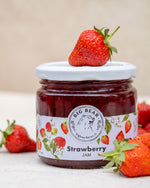A jar of Big Bear Farms Strawberry Jam on a light surface with whole strawberries around it. The jar has a white lid and a white label with red strawberries and the Big Bear logo.