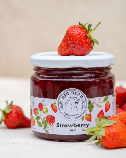 A jar of Big Bear Farms Strawberry Jam on a light surface with whole strawberries around it. The jar has a white lid and a white label with red strawberries and the Big Bear logo.