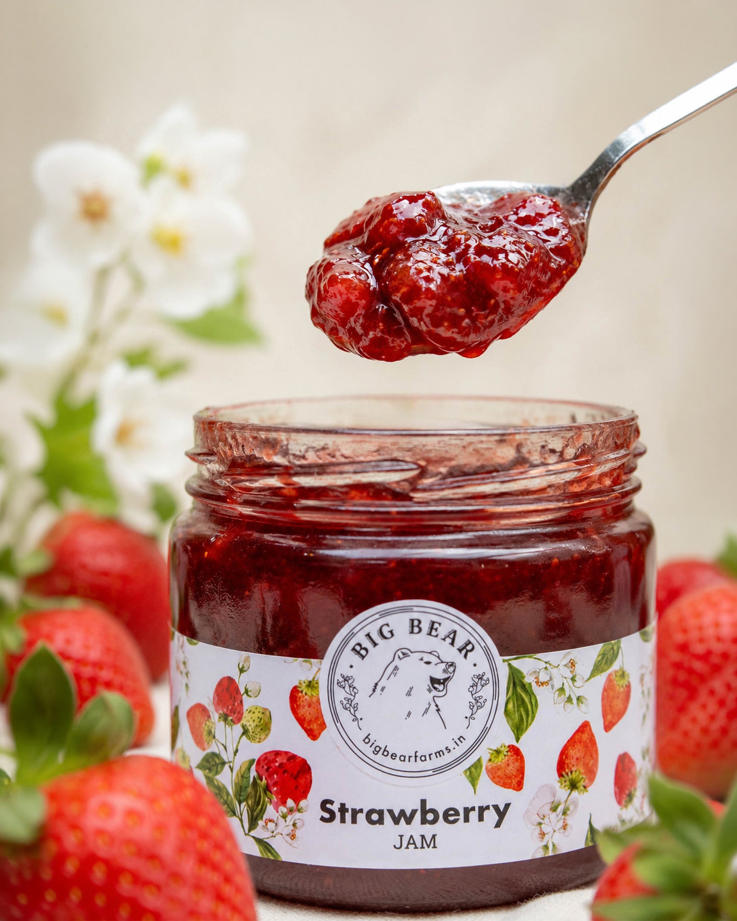 A glass jar of Big Bear Farms Strawberry Jam with a spoonful of glossy jam above the open jar, surrounded by fresh strawberries and soft white flowers in a warm, blurred setting.