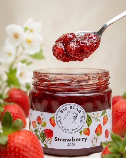 A glass jar of Big Bear Farms Strawberry Jam with a spoonful of glossy jam above the open jar, surrounded by fresh strawberries and soft white flowers in a warm, blurred setting.