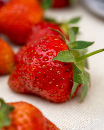 A close-up shot of a fresh Big Bear Farms Strawberries, ripe red strawberry with green leaves on a light beige textured fabric background, with other strawberries blurred in the background.