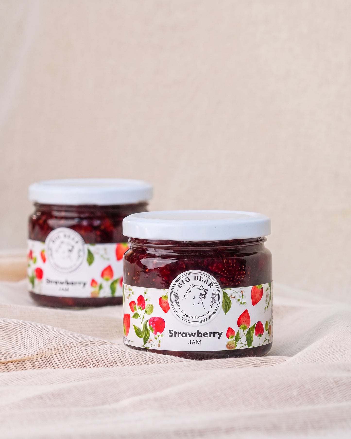 Two glass jars of Big Bear Farms Strawberry Jam arranged on a draped beige fabric surface against a neutral beige background.