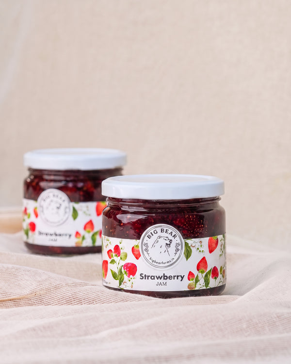 Two glass jars of Big Bear Farms Strawberry Jam arranged on a draped beige fabric surface against a neutral beige background.