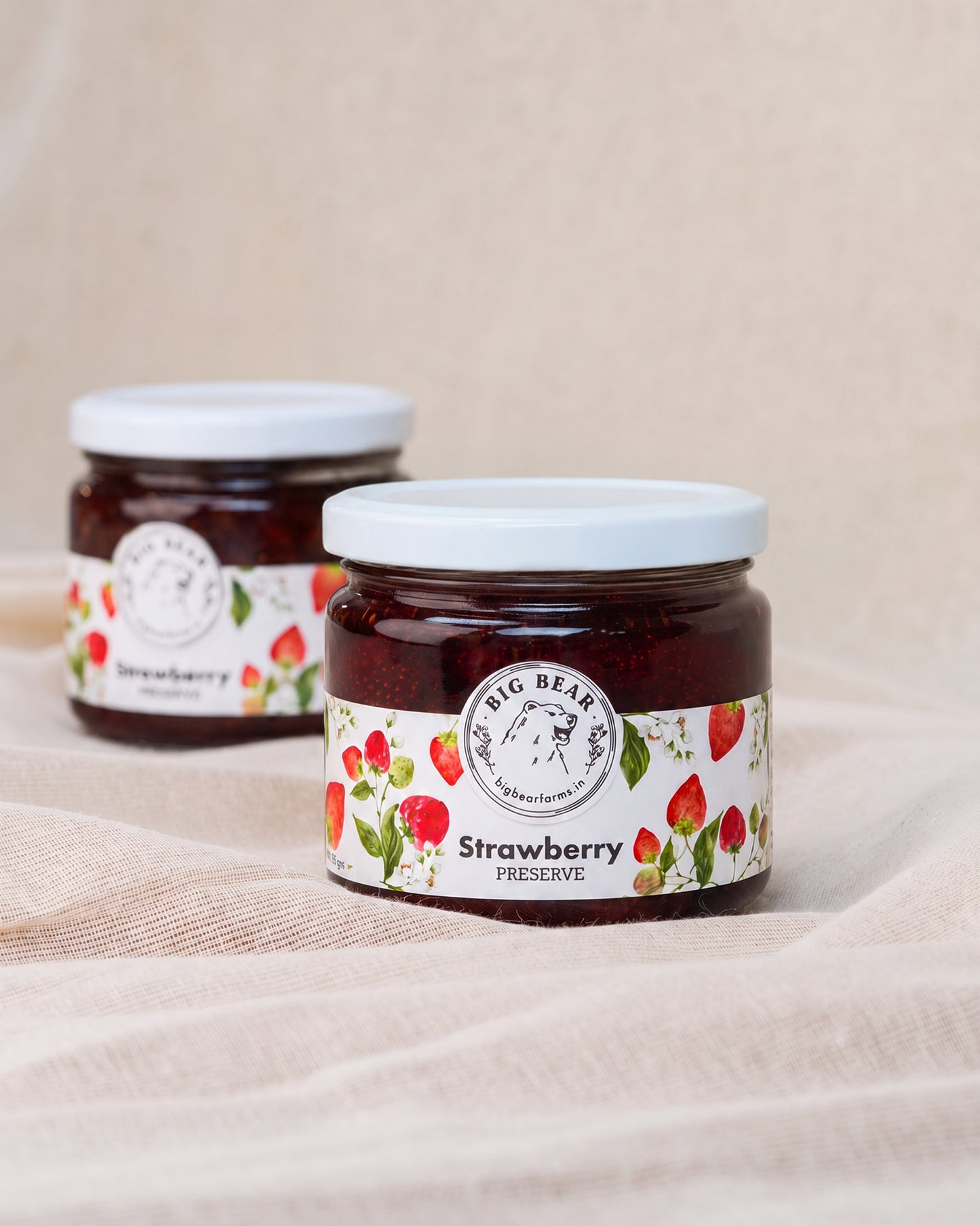 Two glass jars of Big Bear Farms Strawberry preserve with white lids and white labels decorated with strawberries, set on a beige fabric background.
