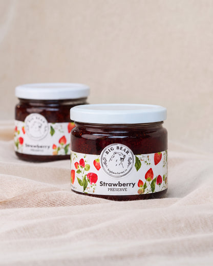 Two glass jars of Big Bear Farms Strawberry preserve with white lids and white labels decorated with strawberries, set on a beige fabric background.