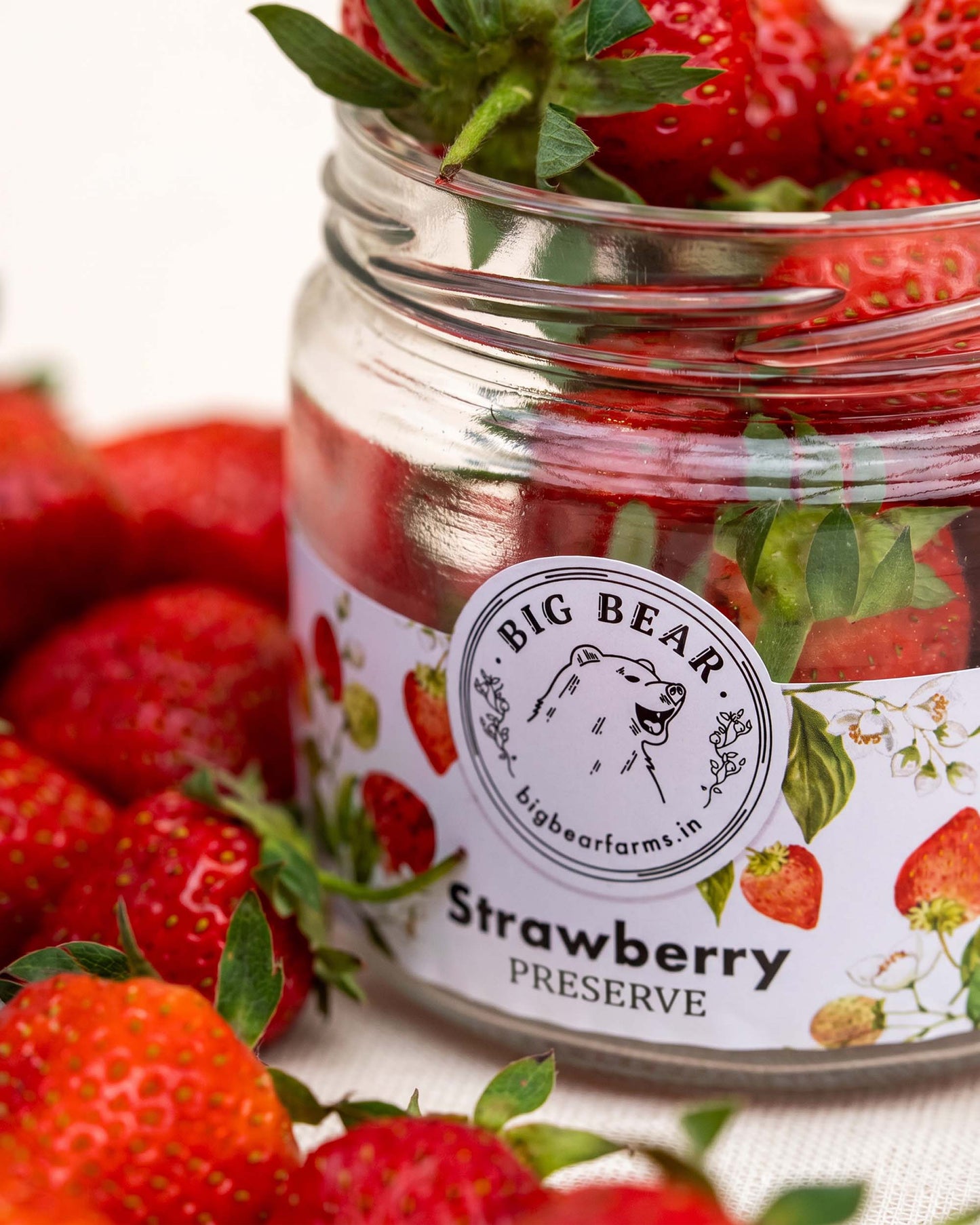 A glass jar of Big Bear Farms Strawberry Preserve filled with red strawberries and topped with fresh green leaves, surrounded by more strawberries on a light fabric background.