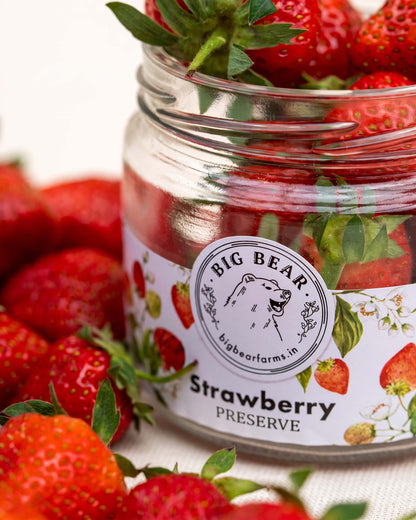 A glass jar of Big Bear Farms Strawberry Preserve filled with red strawberries and topped with fresh green leaves, surrounded by more strawberries on a light fabric background.