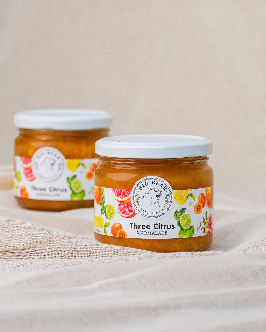 Two glass jars of Big Bear Farms Three Citrus Marmalade with white lids, set on a draped beige linen fabric background.