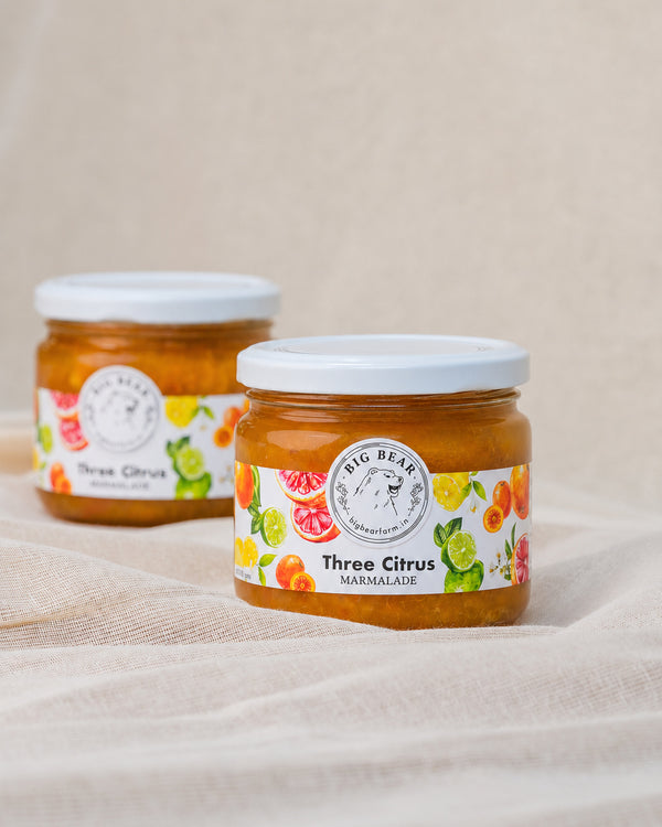 Two glass jars of Big Bear Farms Three Citrus Marmalade with white lids, set on a draped beige linen fabric background.