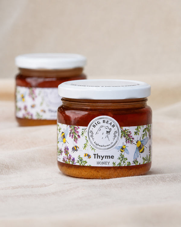 Two glass jars of Big Bear Farms Thyme Honey arranged on a draped beige fabric background, with the front jar in sharp focus and the rear jar softly blurred.