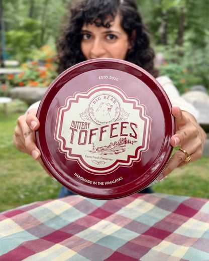 A person outdoors holds up a round dark red metal tin of 40 pieces Big Bear Farms Buttery Toffees in front of their face. The background shows a blurred green garden setting, and the person is standing behind a red, white, and blue plaid tablecloth.