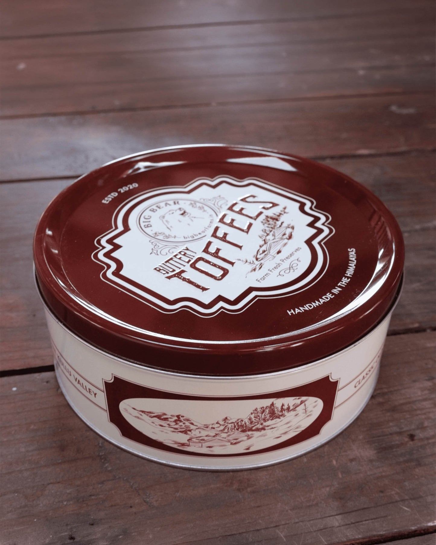 A round metal tin of 40 Big Bear Farms Buttery Caramel Toffees on a rustic dark wood plank surface. The tin has a dark brown lid with a cream-colored vintage label and a matching cream-colored lower body with brown illustrations.