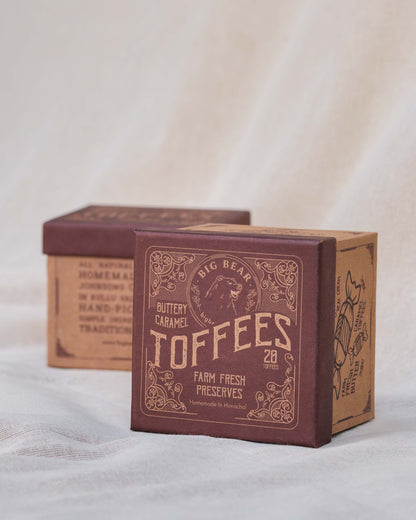 A square box of 20 Big Bear Farms Buttery Caramel Toffees, with the front box showing a dark maroon label with cream text and illustrations, and a second box in the background on a soft beige fabric surface.