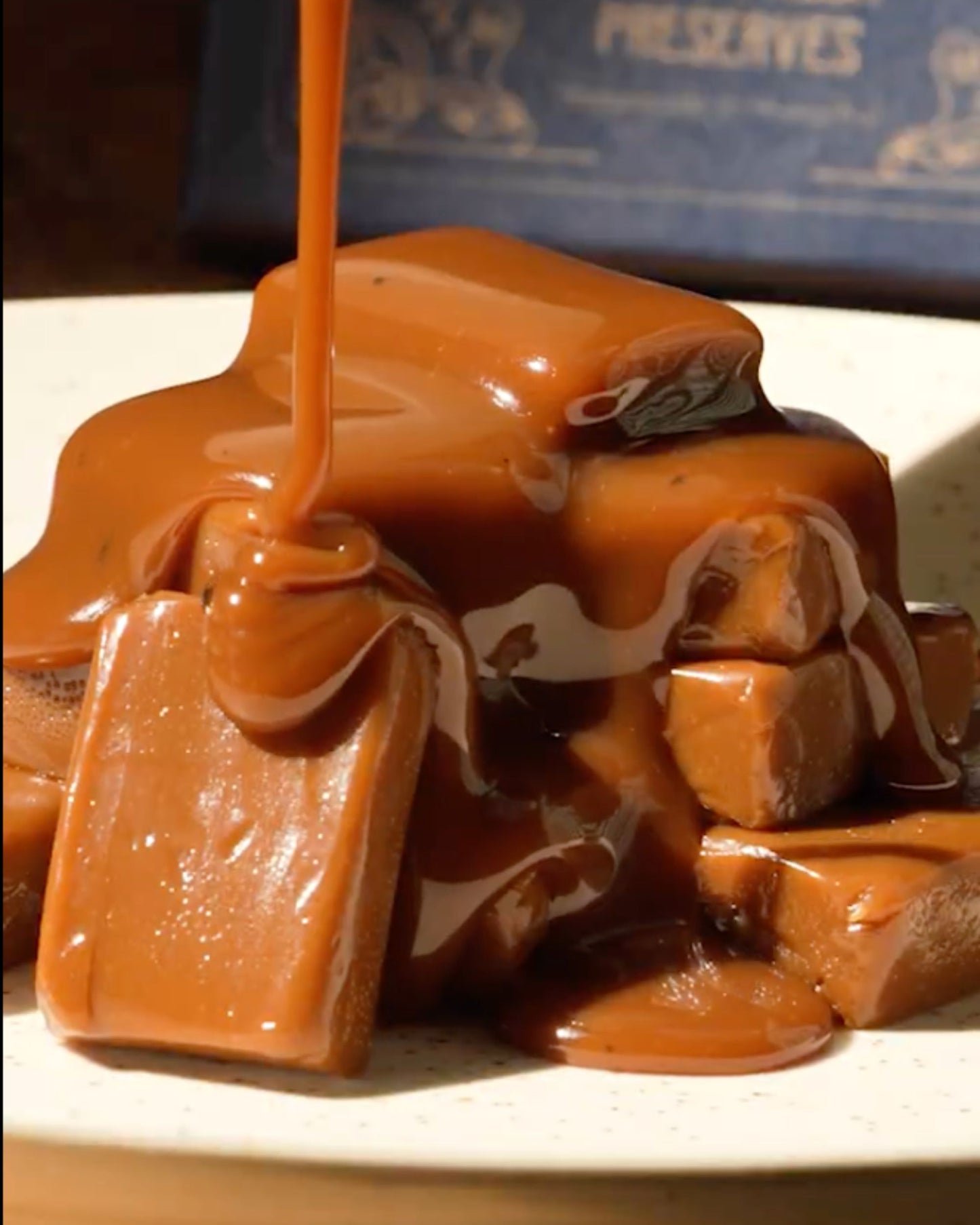 A close-up of Big Bear Farms buttery caramel-topped toffee pieces on a plate, with glossy caramel sauce being poured over them. 