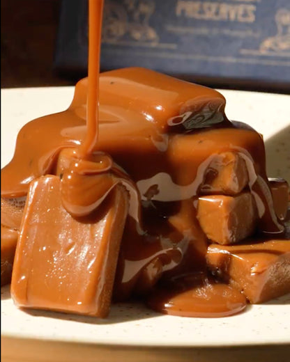 A close-up of Big Bear Farms buttery caramel-topped toffee pieces on a plate, with glossy caramel sauce being poured over them. 