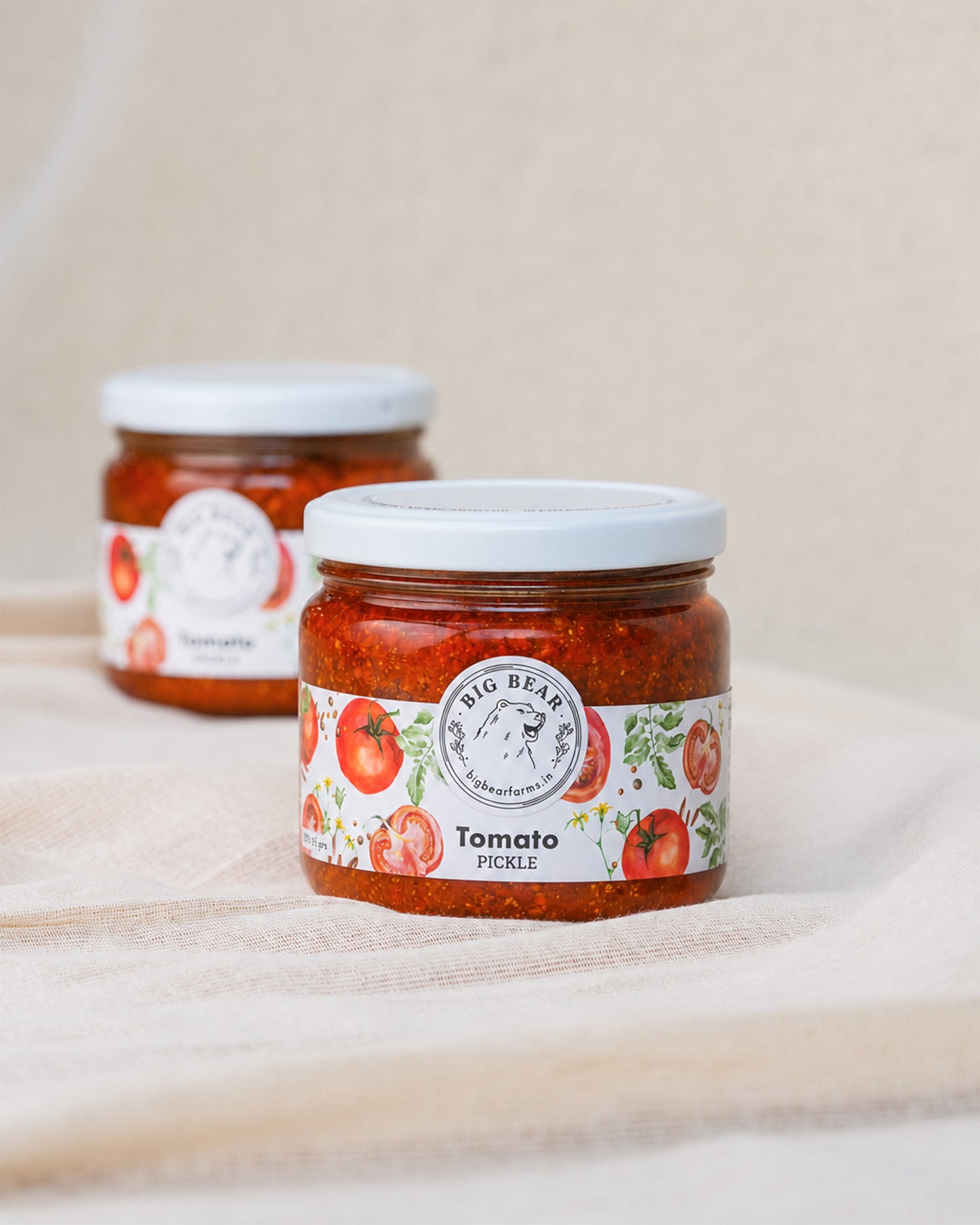 Two glass jars of Big Bear Farms Tomato Pickle, with one jar in sharp focus in the foreground and the other blurred in the background, set on a beige fabric surface.