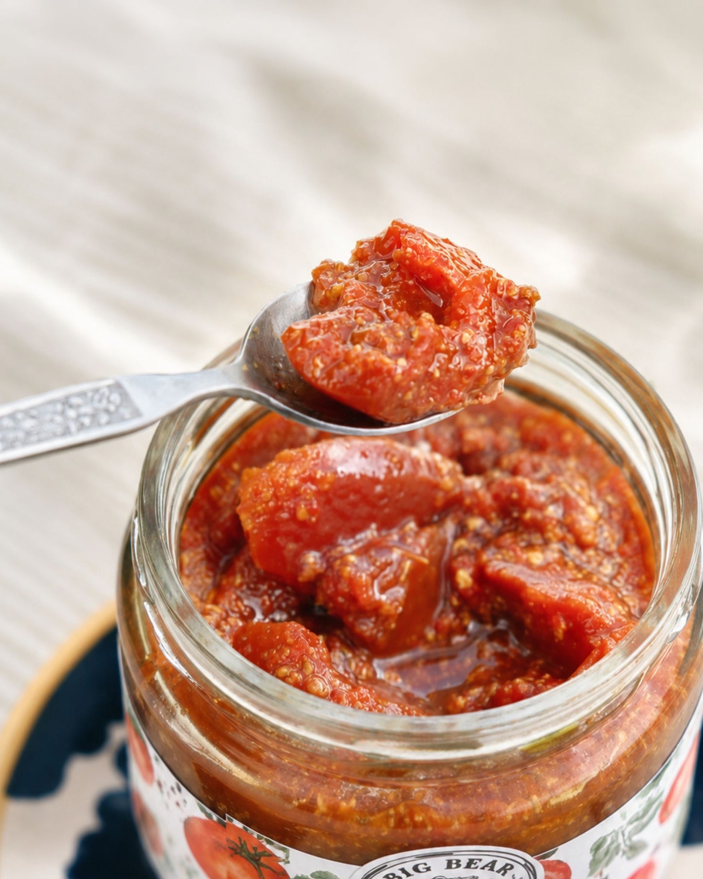 A close-up of a glass jar filled with chunky Big Bear Farms Tomato Pickle , with a spoon lifting a scoop of the thick textured Big Bear Farms Tomato Pickle . The jar label shows the brand Big Bear.