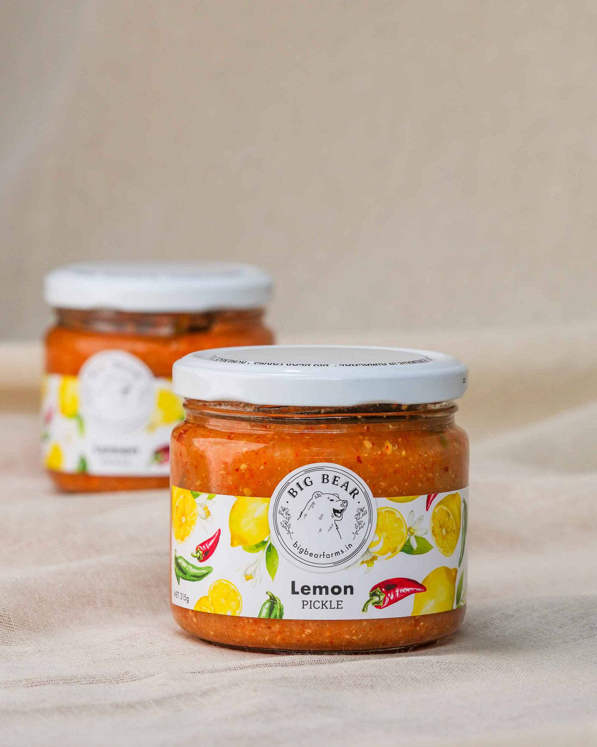 Two glass jars of Big Bear Farms Homemade lemon pickle (nimbu achar) arranged on a beige fabric surface, with the front jar in sharp focus and the rear jar softly blurred in the background.