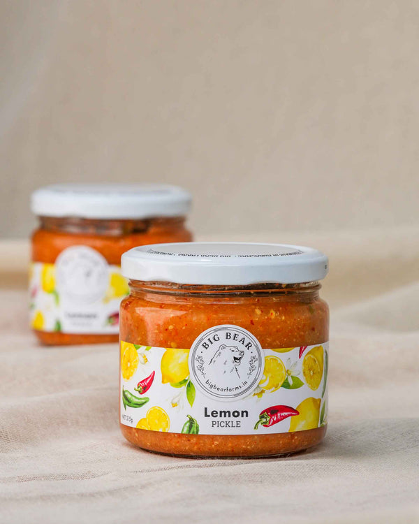 Two glass jars of Big Bear Farms Homemade lemon pickle (nimbu achar) arranged on a beige fabric surface, with the front jar in sharp focus and the rear jar softly blurred in the background.